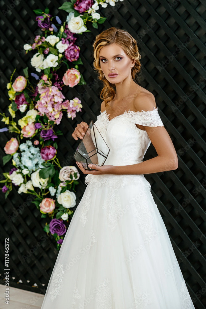 Beautiful girl blonde bride with hair and makeup posing in a wedding dress in the dark interior, flowers in the background. Beautiful girl blonde bride with hair and makeup posing in a wedding dress in the dark interior, flowers in the background.