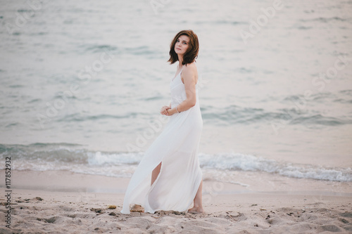 Young beautiful woman in a white dress walking on an empty beach near ocean