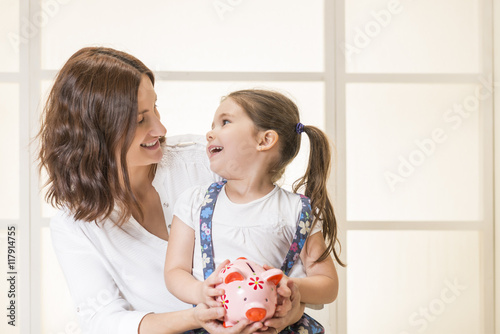 Happy mother offering piggy bank to her beautiful daughter. Family budget and savings concept. Education, school and money saving concept. Junior Savings Account concept