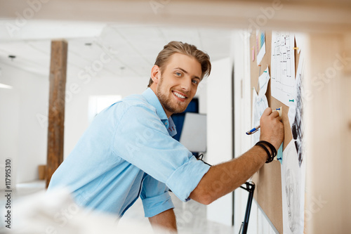 Young handsome businessman writing on the paper pinned to corkboard, looking in camera smiling. Office background.