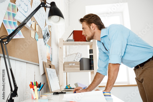 Young handsome businessman working standing at table and looking thruogh papers pinned to corkboard. Office background. photo