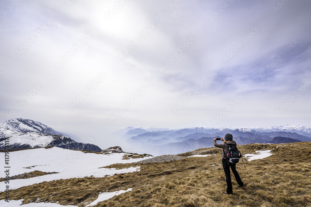 Fototapeta premium Girl taking a picture of mountain landscape in the middle of a field with some snow at Monte Altissimo, San Giacomo, Trentino region, Italy