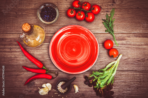 Fresh tomatoes, chili pepper and other spices and herbs around modern red plate in the center of wooden table and cloth napkin. Top view. Blank place for your text. Close-up.