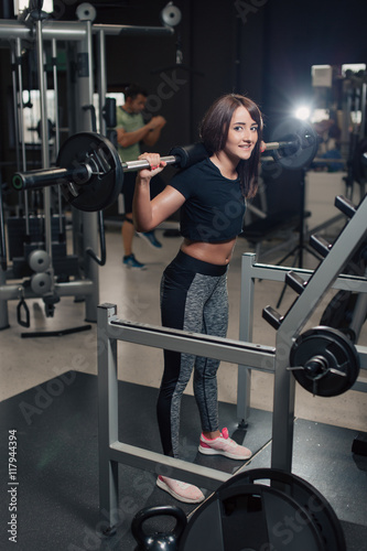 Young strong woman doing squats at the gym with a barbell on her back while smiling at the camera and other people in the background 