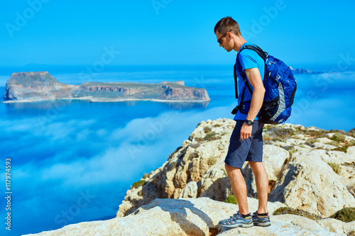 male traveler with backpack standing on the trail on the cliff against sea and blue sky at early morning. Balos beach on background, Crete, Greece