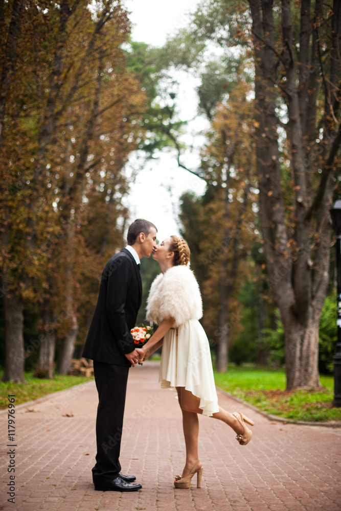 Stylish bride in beige dress kisses a groom on the path in park Stylish bride in beige dress kisses a groom on the path in park