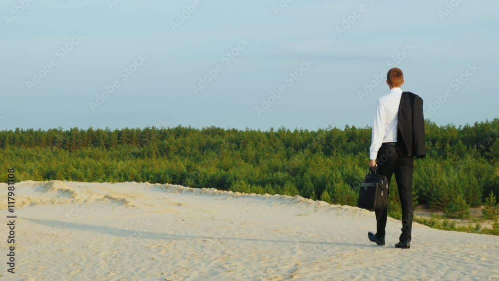 Carefree businessman walking on the sand, carries a jacket over his shoulder. Back view Carefree businessman walking on the sand, carries a jacket over his shoulder. Back view