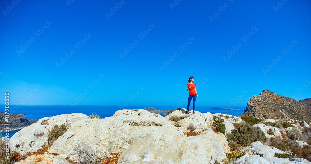 sporty female traveler with backpack standing on the cliff against sea and blue sky with white clouds at early morning, Crete, Greece.