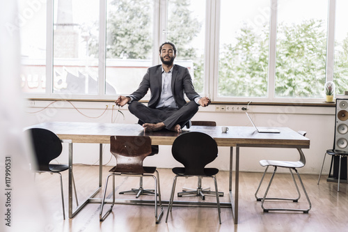 Young businessman sitting cross-legged on desk in office