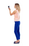 back view of standing young beautiful  woman  and using a mobile phone. girl  watching. Rear view people collection.  backside view of person.  Isolated over white background. Blonde in blue pants