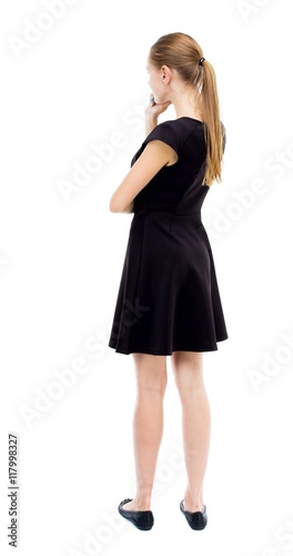 back view of standing young beautiful  woman.  girl  watching. Rear view people collection.  backside view of person.  The blonde in a black dress thoughtfully put a hand to his chin.