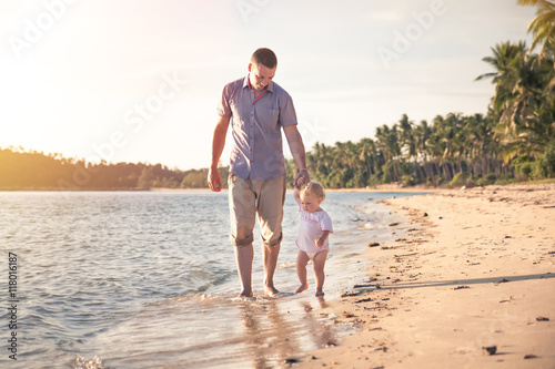Father and child walking together on the beach coast during sunset with shining sea on background. Father holding child hand © splendens