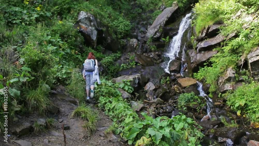 Hiker woman with backpack walking on a forest trail in the mountains on the background a waterfall. Hiking. Adventure in the forest. Hiker woman with backpack walking on a forest trail in the mountains on the background a waterfall. Hiking. Adventure in the forest.