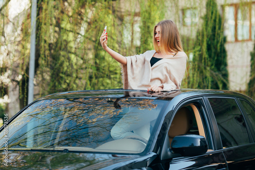 Picture of cheerful young woman wearing sunglasses and raised hands on the sunroof of the luxury car photo