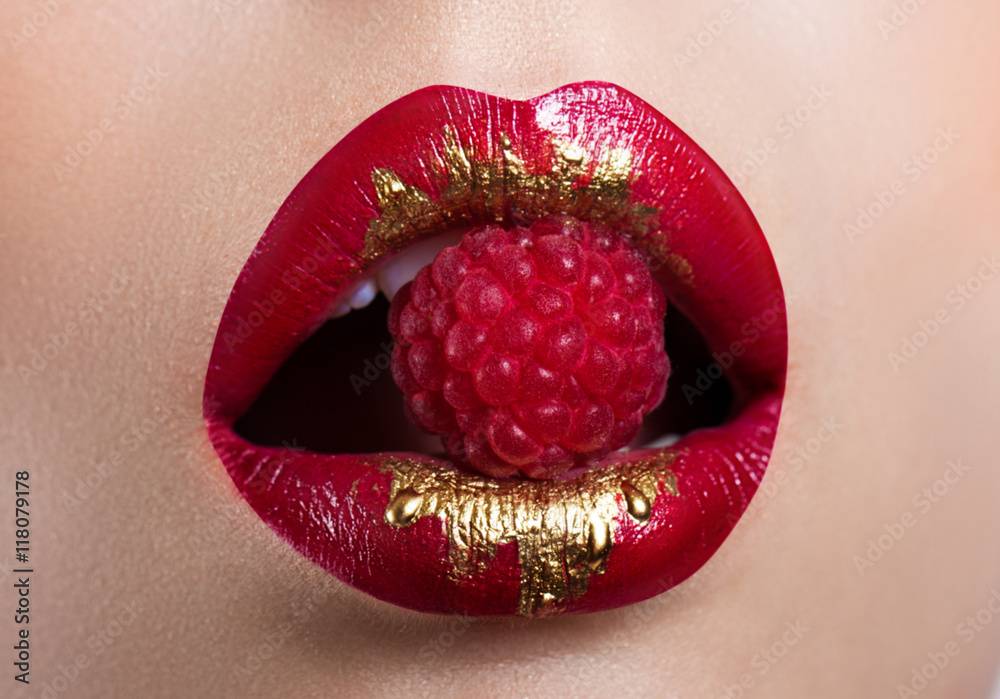 Fototapeta premium Beautiful female lips closeup. Red lipstick with gold. Berry raspberry in mouth
