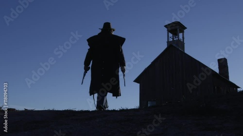 Silhouette of a cowboy with a gun in each hand, walking to a building. 