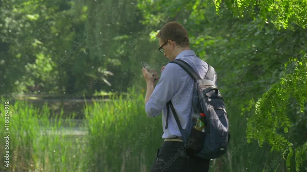 Man Clicks a Screen of Smartphone in Park Holding a Mobile Phone Turns Around Backpacker is Walking at the Nature Fresh Green Trees Sunny Summer Day Man Clicks a Screen of Smartphone in Park Holding a Mobile Phone Turns Around Backpacker is Walking at the Nature Fresh Green Trees Sunny Summer Day