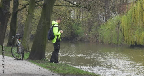 Man is Looking at River Clicks a Tablet Bicycle is Placed at the Tree Riding a Bicycle in Park Alley Backpacker Near the River Water Flows Spring Cloudy photo