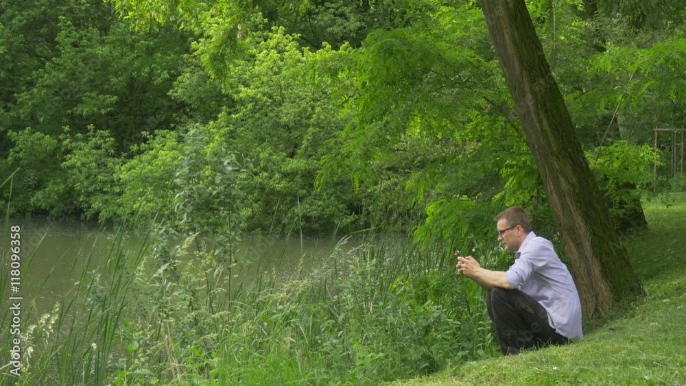 Man Clicks Mobile Phone Lake Bank Man is Texting Chatting Sitting Near the Water Under Acacia Tree Holding a Smartphone Fresh Green Trees Sunny Summer Day