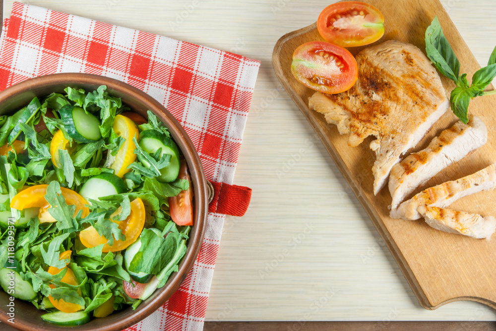 Chicken fillet, grilled, with a large portion of salad with fresh vegetables and herbs on a wooden table with a red cloth into a cell. top view