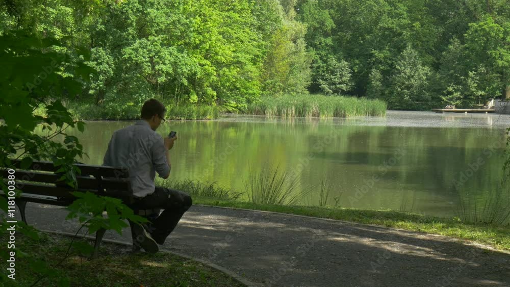 Man Sitting on a Bench Talking on Smartphone Lake at Green Park Man Holding Mobile Phone Stands up Walking Away Trees Grow Along the Alley Reed in Water Man Sitting on a Bench Talking on Smartphone Lake at Green Park Man Holding Mobile Phone Stands up Walking Away Trees Grow Along the Alley Reed in Water
