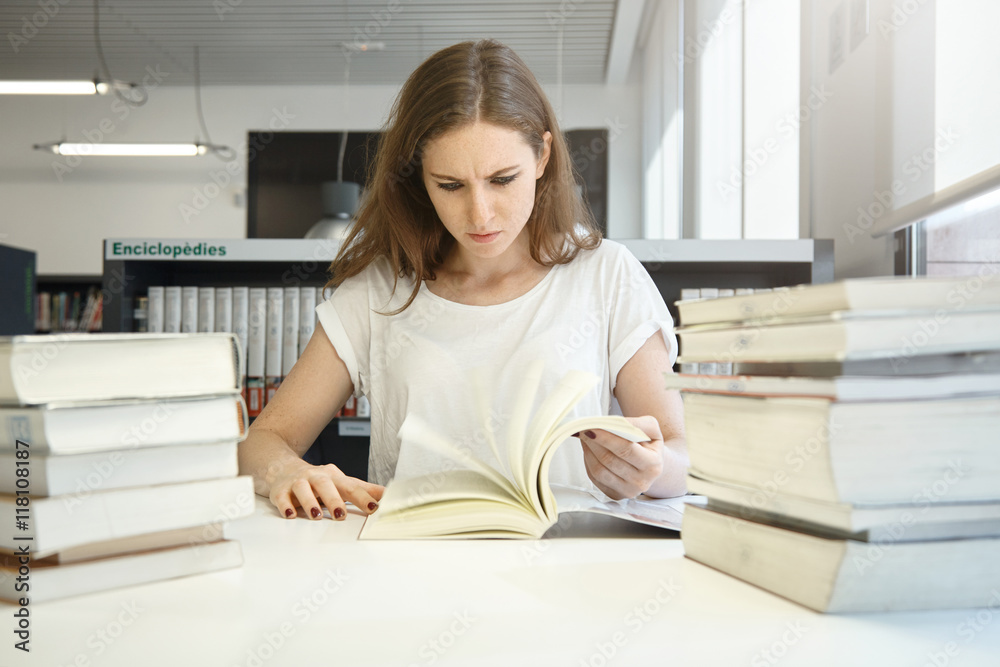 Human emotions and feelings. Stressed student girl preparing for final exams, studying at library in front of big piles of books, staring at textbook, looking shocked with how much work she has to do Human emotions and feelings. Stressed student girl preparing for final exams, studying at library in front of big piles of books, staring at textbook, looking shocked with how much work she has to do