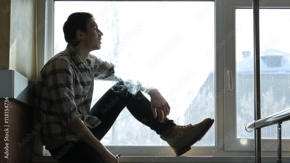 Handsome man sits on windowsill and smokes electronic hookah.