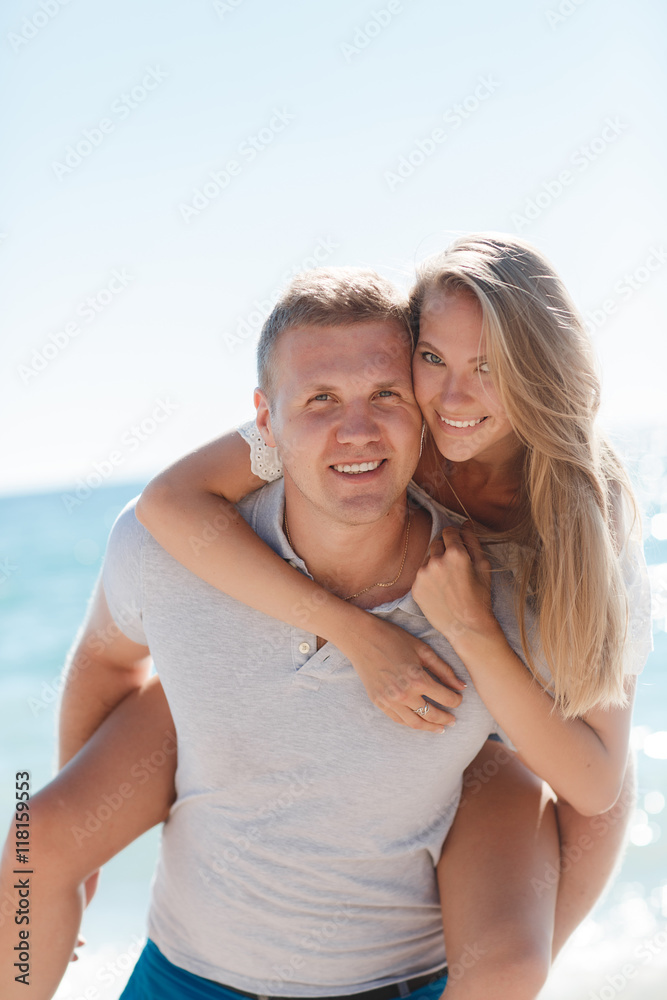 Happy loving couple,young blond woman having fun on the beach near the blue ocean,a girl is sitting on the back of a guy hugging his neck the girl wears a ring on his right hand,he wears a gold chain Happy loving couple,young blond woman having fun on the beach near the blue ocean,a girl is sitting on the back of a guy hugging his neck the girl wears a ring on his right hand,he wears a gold chain