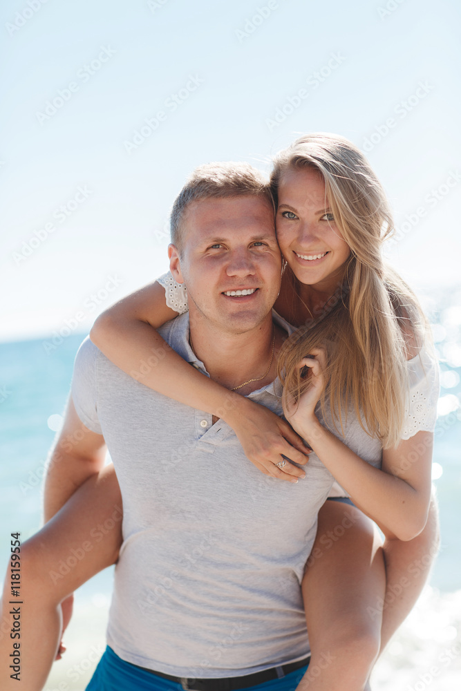 Obraz premium Happy loving couple,young blond woman having fun on the beach near the blue ocean,a girl is sitting on the back of a guy hugging his neck the girl wears a ring on his right hand,he wears a gold chain