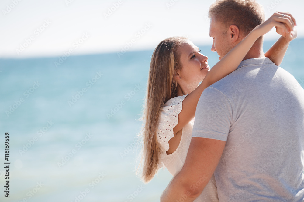 Fototapeta premium Happy young couple blond man and woman spend time together on the beach on the shore of the blue ocean,standing on the sand in an embrace on a background of calm sea and clear sky