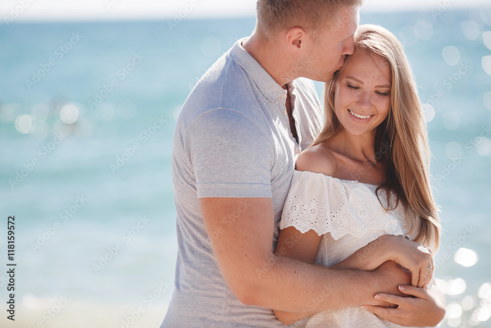 Happy young couple blond man and woman spend time together on the beach on the shore of the blue ocean,standing on the sand in an embrace on a background of calm sea and clear sky Happy young couple blond man and woman spend time together on the beach on the shore of the blue ocean,standing on the sand in an embrace on a background of calm sea and clear sky