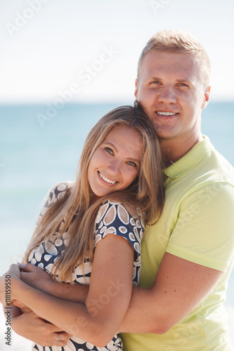 Happy young couple blond man and woman spend time together on the beach on the shore of the blue ocean,standing on the sand in an embrace on a background of calm sea and clear sky © GTeam