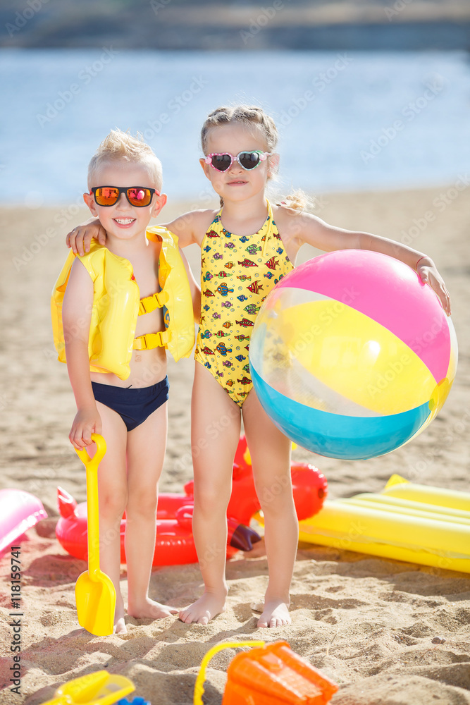 Little girl with pigtails and a boy with short hair girl,dressed in a  yellow swimsuit,a boy dressed in a yellow inflatable lifejacket,standing on  the beach with multi-colored inflatable ball Stock Photo |