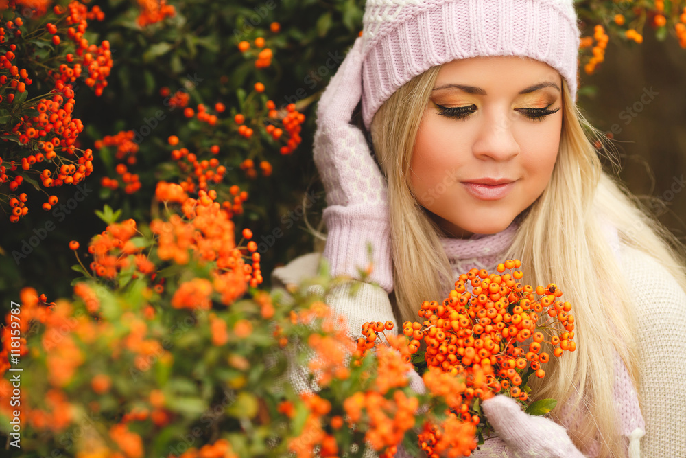 Naklejka premium young woman with long blonde straight hair,wearing a light sweater,pink scarf and gloves,on his head wearing a pink and white knitted cap,spends time outdoors in the Park near bushes of ripe red Rowan