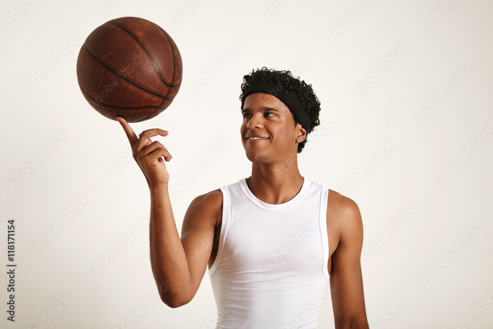 Closeup shot of a playful smiling young attractive African American basketball player wearing a white sleeveless shirt holding an old leather ball on one finger isolated on white.