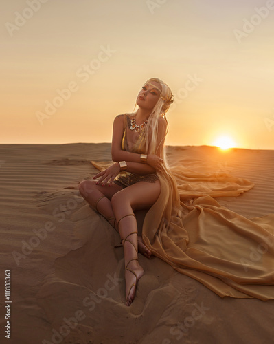 Beautiful queen of the desert sitting on the sand. Luxury golden dress and unusual jewelry. Gold flower in her hair, hairstyle with braids. Beautiful blonde. Fashionable toning.