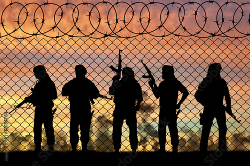 Silhouette of terrorists with rifles standing against fence of barbed wire during sunset