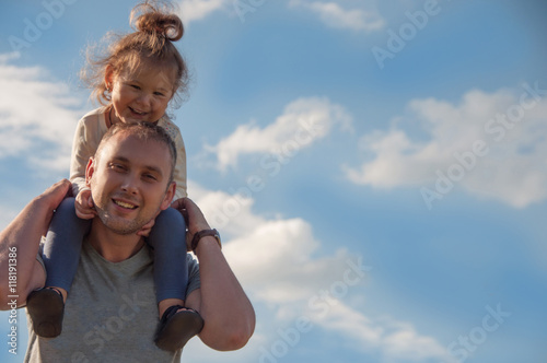 Happy daughter on his shoulders at father against the sky