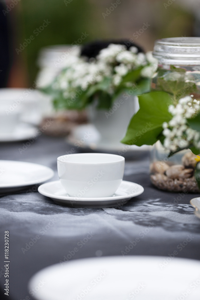 Modestly decorated wedding table. Gothic romance, garden wedding. White cups standing on a black tablecloth with handmade white pattern. Clean light, white plates, handcrafted decoration, black roses.