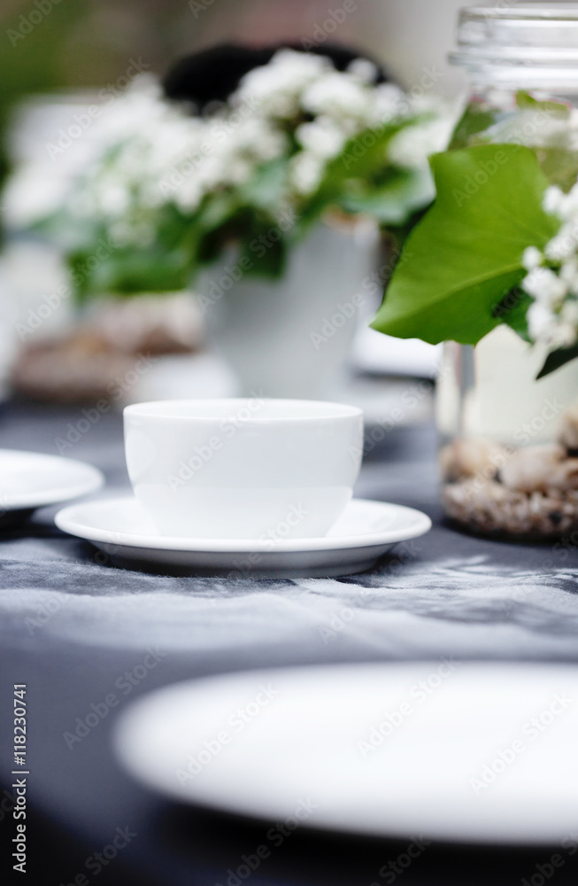 Modestly decorated wedding table. Gothic romance, garden wedding. White cups standing on a black tablecloth with handmade white pattern. Clean light, white plates, handcrafted decoration, black roses.