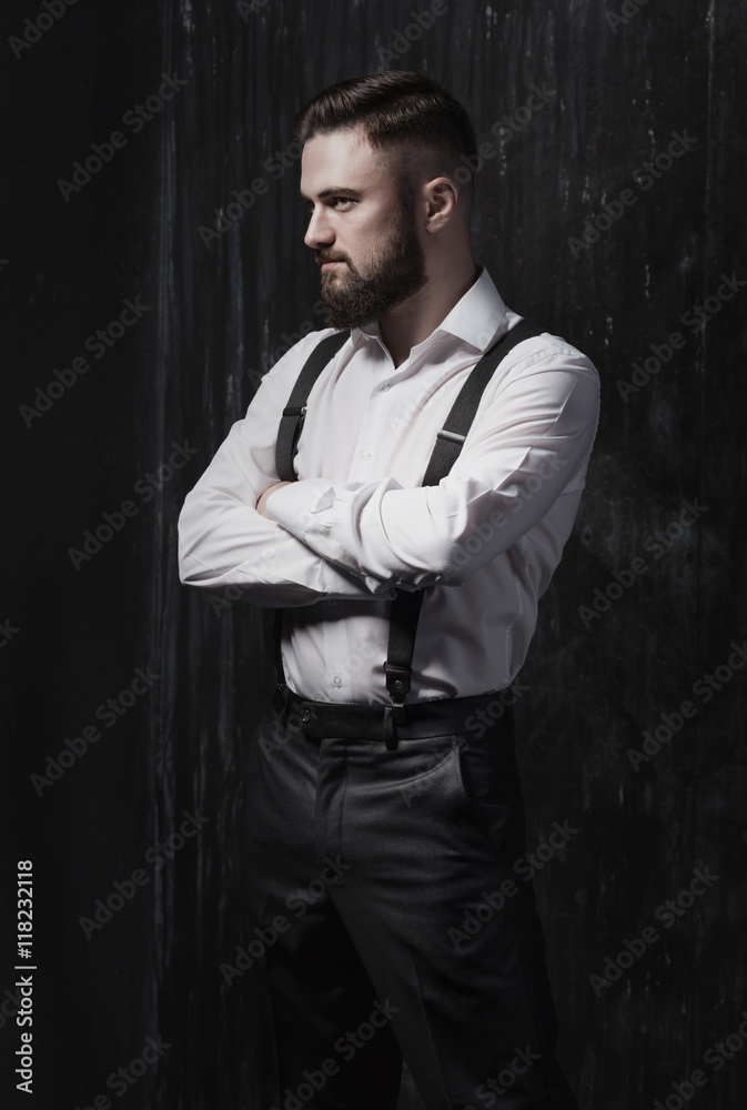 Handsome bearded man in a white shirt and suspenders standing near dark wall. He is strong, courageous and serious. Dark room, night and harsh shadows.