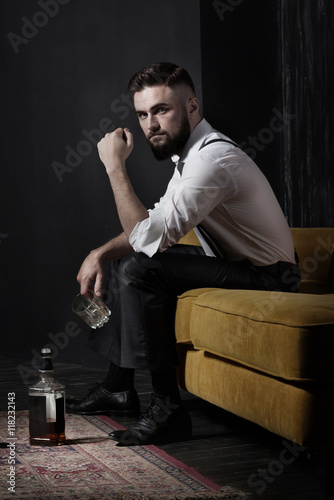 A handsome brutal bearded man sitting on an orange couch. He is holding an empty glass. Whiskey bottle standing on the floor. He looks at the viewer. Dark room, old carpet, deep shadows.