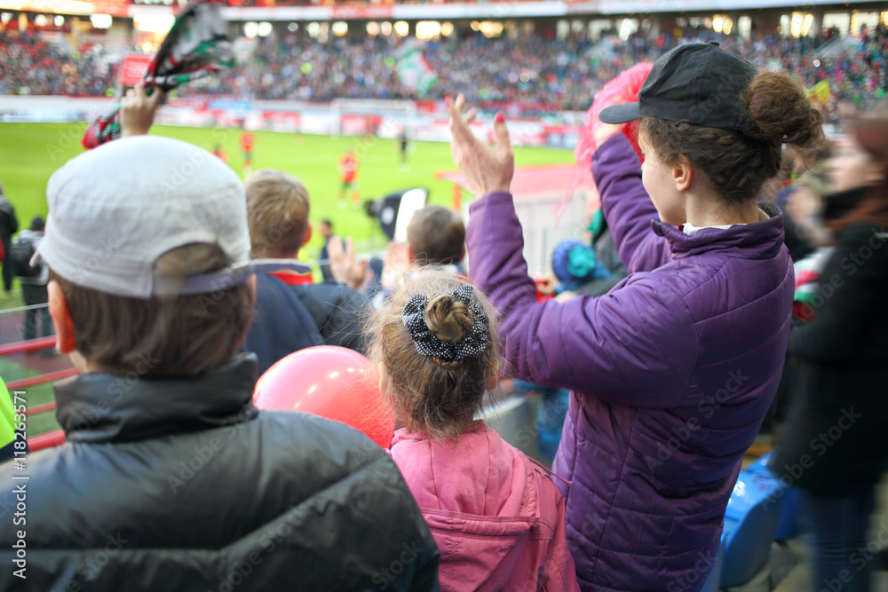 Woman, girl and boy standing applause among fans at a sports stadium, the view from the back