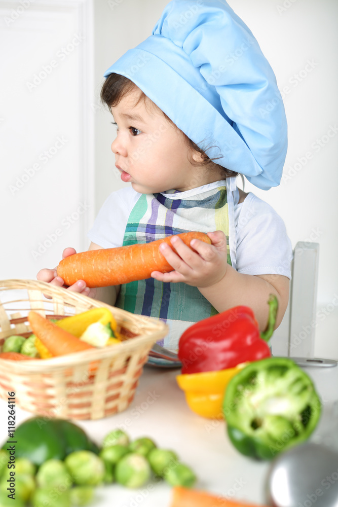 Little boy with serious face in kitchen apron and cap holding carrot Little boy with serious face in kitchen apron and cap holding carrot