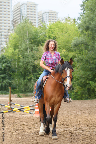 Happy woman with curly hair riding a horse in park near the apartment complex © Pavel Losevsky