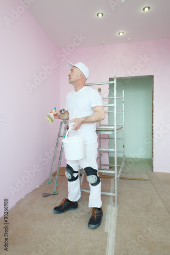 Plasterer in white clothes and kneepads looking at his work in pink room photo