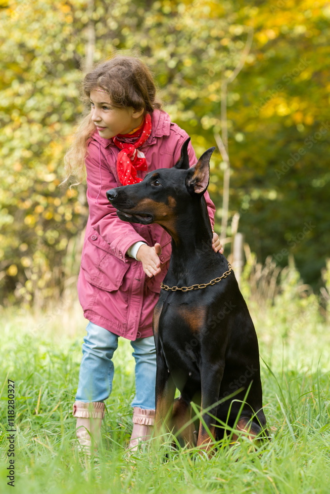 Little curly girl and dobermann are looking to the right in park, focus on a dog. Little curly girl and dobermann are looking to the right in park, focus on a dog.