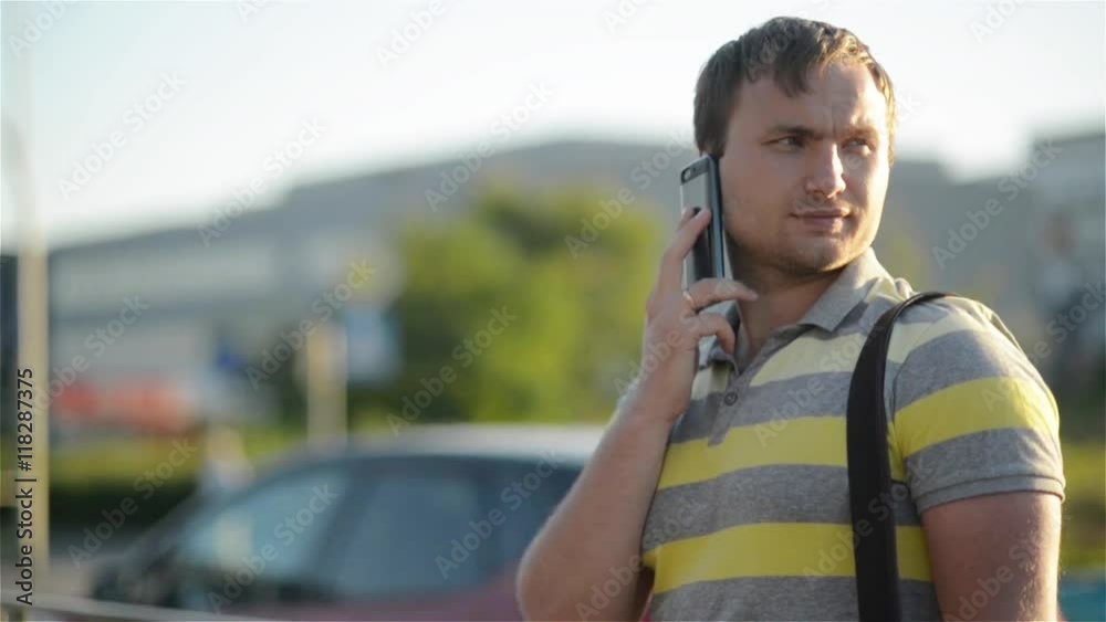 Young man holding mobile phone, using smartphone, making a call, talking on the phone, standing on sunny street with transport traffic on the background Young man holding mobile phone, using smartphone, making a call, talking on the phone, standing on sunny street with transport traffic on the background