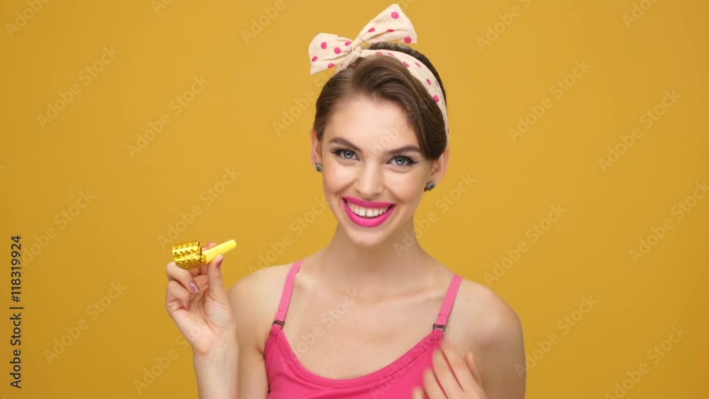 Amusing cute young pinup woman in yellow dress blowing in party whistle and laughing Amusing cute young pinup woman in yellow dress blowing in party whistle and laughing
