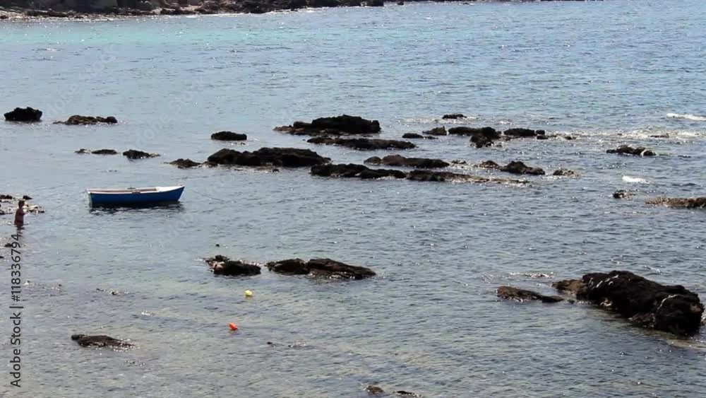 A distant figure wades in the waters of the Mediterranean Sea near rocks and a boat. A distant figure wades in the waters of the Mediterranean Sea near rocks and a boat.